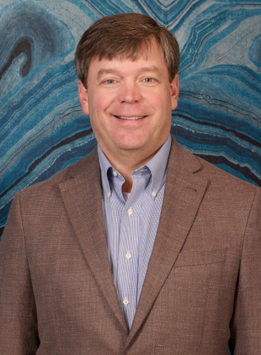 Man in a brown blazer and blue shirt smiling, standing in front of a blue marbled background.