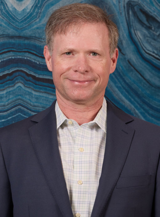 Smiling person in a suit with a blue and white checkered shirt in front of a blue patterned background.