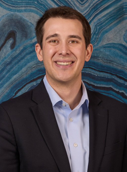 A person in a suit and light blue shirt smiles in front of a blue marbled background.