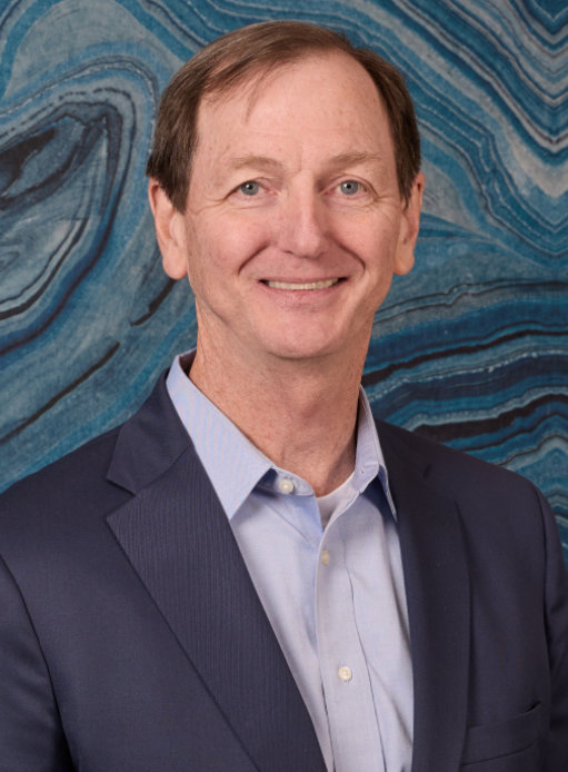 A man in a suit and light blue shirt smiles in front of a blue and black marbled background.