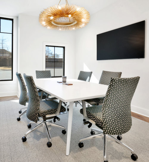 A modern conference room with a white table, six patterned chairs, a large wall-mounted screen, and a decorative chandelier. Natural light enters through two windows.