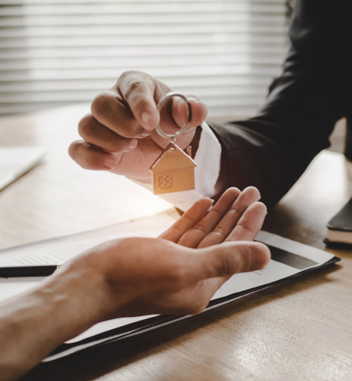 Hand gives house-shaped keychain to another hand over a desk with documents.