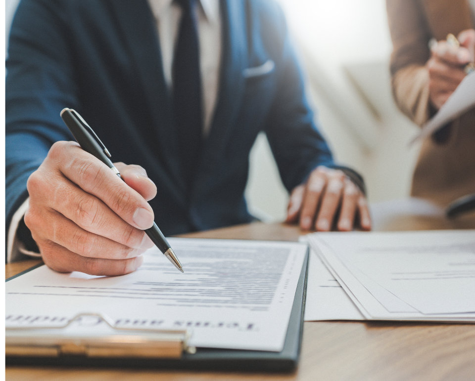 Person in a suit signing a contract on a clipboard, with another person standing nearby, holding documents.